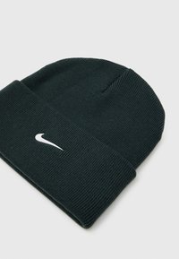 Tamna zelena pletena kapa s preklopom, s bijelim Nike logom sprijeda. Izrađena od mekog, teksturiranog materijala.