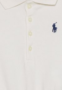 Valkoinen polo-paita, jossa on kaulus, neljä nappia ja pieni sininen brodeerattu polo-pelaajan logo rinnassa.