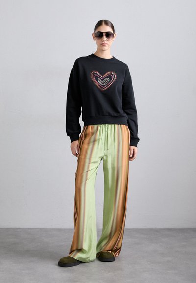 Črn pulover z večbarvnim srčnim vzorcem, v kombinaciji z zelenimi satenskimi hlačami, ki imajo rjave stripe. Model nosi sončna očala in natikače.