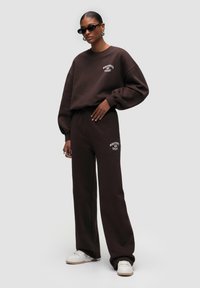 Brauner Baumwoll-Sweatshirt in oversized Passform mit weißem Logo, kombiniert mit passenden braunen Jogginghosen. Weiße Sneakers runden den Look ab.