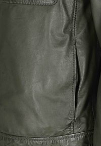 Olivegrüne Lederjacke mit weicher Textur, ausgestattet mit einer Fronttasche und seitlichem Reißverschlussdetail, das das glatte und strapazierfähige Material betont.