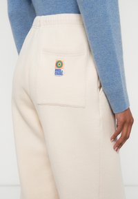 Béžové tepláky s mäkkou textúrou, ktoré majú zadnú kapsu a farebnú nášivku s logom "House of Colors". Dopĺňa ich svetlomodrý top.