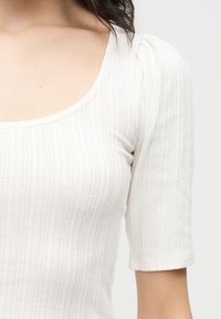 Κοντινό πλάνο ενός ατόμου που φοράει ένα λευκό, ριγωτό, μπλουζάκι με κοντά μανίκια, στρογγυλεμένο λαιμό και φουσκωτές λεπτομέρειες στους ώμους.