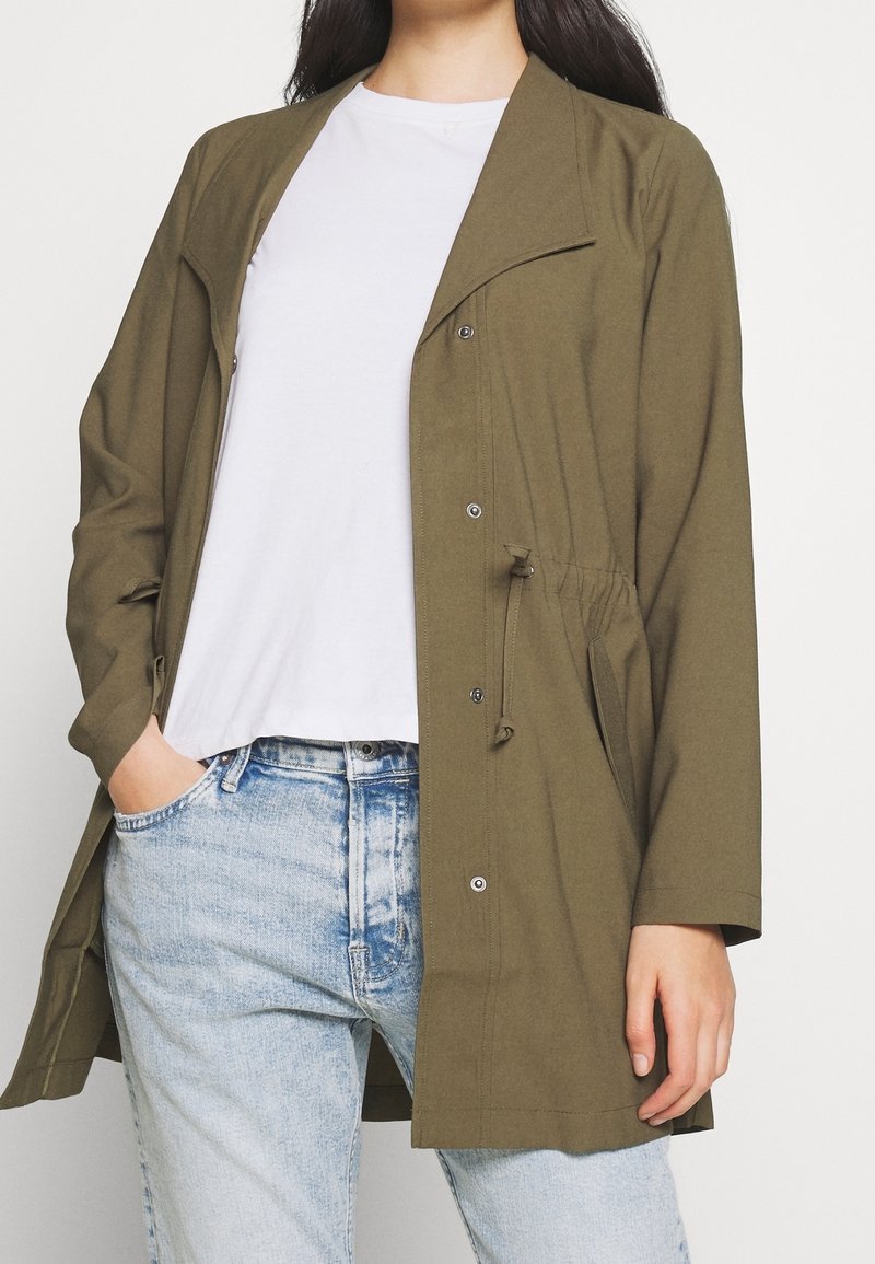 Olive green, lightweight jacket with a relaxed fit, snap front closure, and adjustable drawstring waist. Paired with a white t-shirt and blue jeans.
