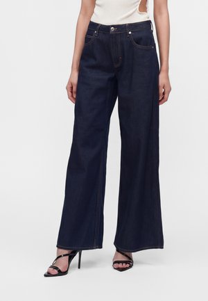 Persona che indossa jeans blu scuro a gamba larga e sandali neri con tacco alto, abbinati a un top bianco senza maniche con lati ritagliati.