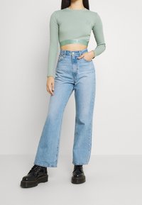Kvinna som bär en salvia grön långärmad croptop, ljusblå högmidjade jeans och svarta kraftiga snörstövlar, stående mot en vit vägg.