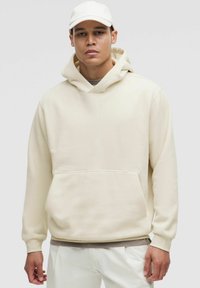 Mand iført en beige hættetrøje og matchende kasket med hvide bukser, der står imod en ensfarvet lys grå baggrund.