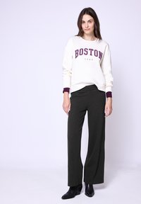 Krémová mikina se sloganem "BOSTON" a rokem "1980" v tmavě červené, spárovaná s tmavě zelenými širokými kalhotami a černými kotníkovými botami.