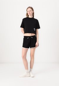 T-shirt nera cropped con logo sul petto, abbinata a pantaloni corti neri con bordi bianchi e una vita con cordino. Sneakers bianche completano il look.