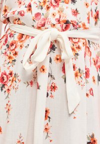 Λευκή υφασμάτινη ζώνη δεμένη σε φιόγκο γύρω από τη μέση με λευκό φόρεμα που έχει κόκκινα και πορτοκαλί λουλουδένια σχέδια.