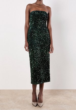 Donna con abito midi senza spalline, di paillettes verde scuro, e décolleté nude a punta, in piedi contro uno sfondo chiaro e semplice.
