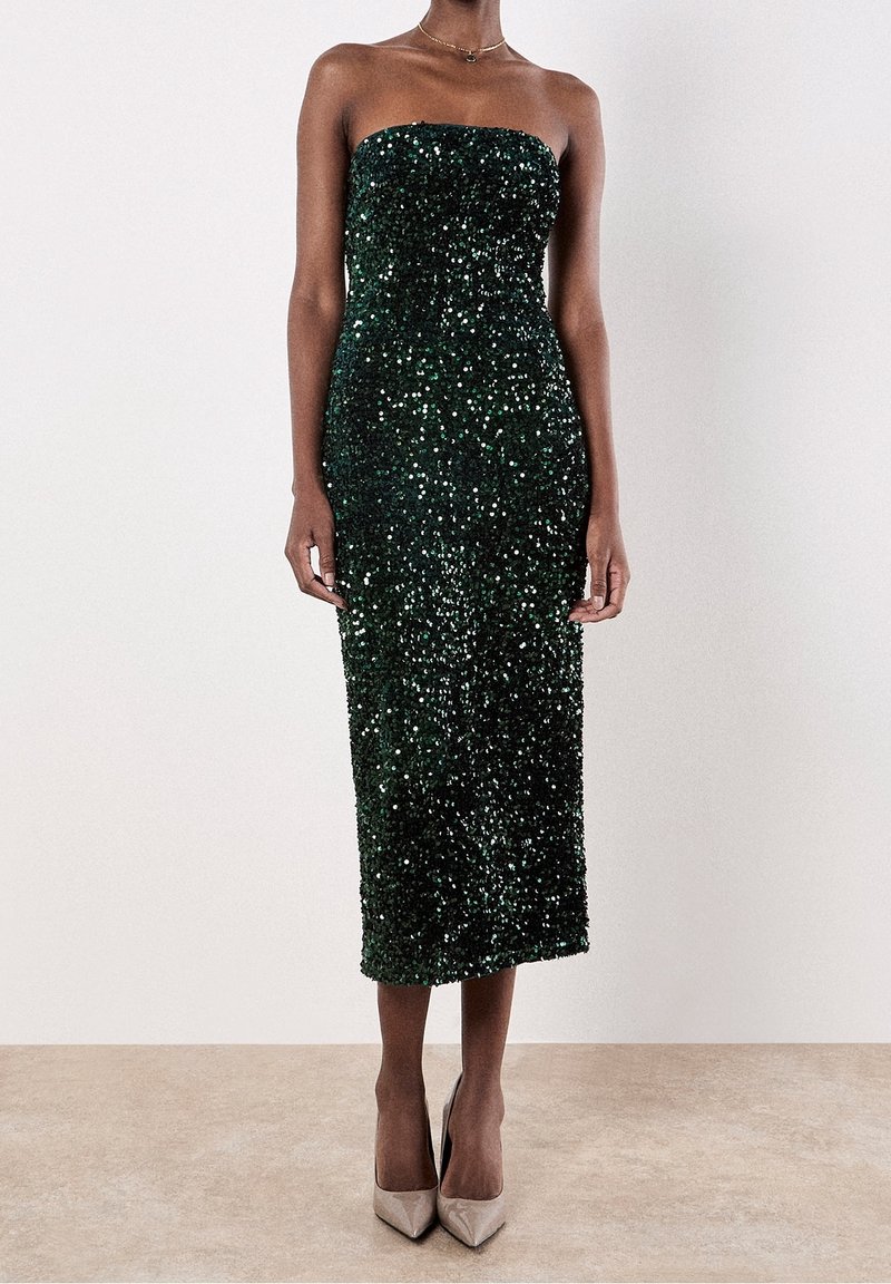 Femme portant une robe midi bustier à sequins vert foncé et des escarpins nude pointus, debout devant un fond uni clair.