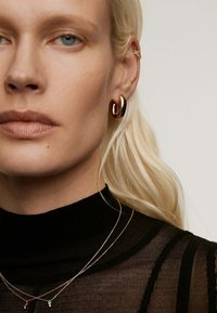 Pendientes de aro dorados, cadenas doradas en capas con acentos de color, top negro de punto transparente, textura de piel suave, cabello largo y rubio.