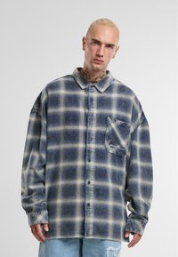 Chemise en flanelle avec un motif à carreaux bleu et crème, design à boutons, coupe oversize, poche poitrine simple et détail brodé.