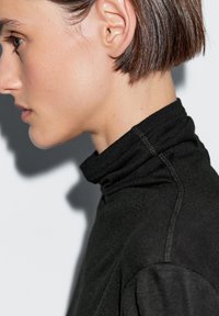 Garment a collo alto di colore nero, con una texture liscia e dettagli di cucitura sottili, indossato da un modello di profilo.