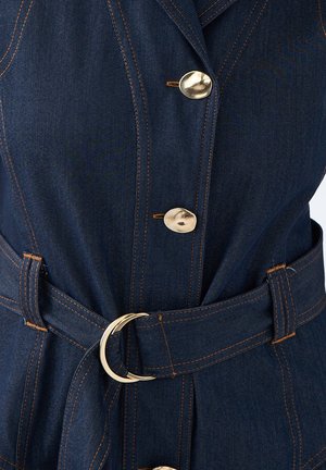 Dunkelblaudress mit goldenen Knöpfen, gesteppten Linien und einem passenden Gürtel mit einer runden Schnalle. Der Stoff hat eine strukturierte Oberfläche.