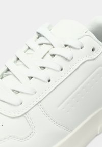 Sneakers en cuir blanc avec lacets plats, col rembourré et semelle en caoutchouc texturé. Présente des perforations subtiles sur les côtés et un design épuré.