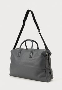 Sac de voyage en cuir gris foncé avec surface texturée, double poignée supérieure, fermeture à glissière et bandoulière noire réglable sur un fond blanc.