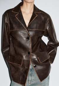 Persoon in een donkerbruin leren blazer met knoopsluiting en klepzakken, gecombineerd met lichtblauwe spijkerbroek, tegen een effen achtergrond.