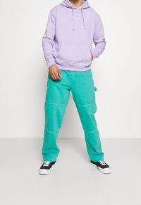 Vaaleanvioletti hupullinen collegepaita etutaskulla, yhdistettynä kirkkaan teal-sävyisiin leveälahkeisiin housuihin ja mustiin skeittikenkiin, joissa on valkoiset pohjat.