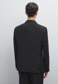 Schwarze maßgeschneiderte Sakko mit schmalen Revers, strukturiertem Stoff und einem Schlitze hinten. Mit knöpfbaren Ärmelabschlüssen.