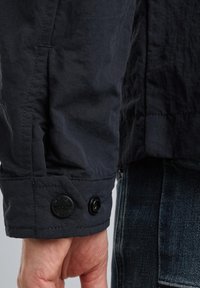 Dunkelblaue Jacke mit strukturierter Oberfläche. Verfügt über einen knöpfbaren Ärmelbund und auffällige Nähte an der Naht. Teilweise sichtbare Jeans aus Denim.