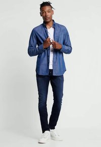 Blauwe denim long-sleeve shirt met een button-down kraag, gecombineerd met een wit T-shirt. Het model draagt donkere jeans en witte sneakers.