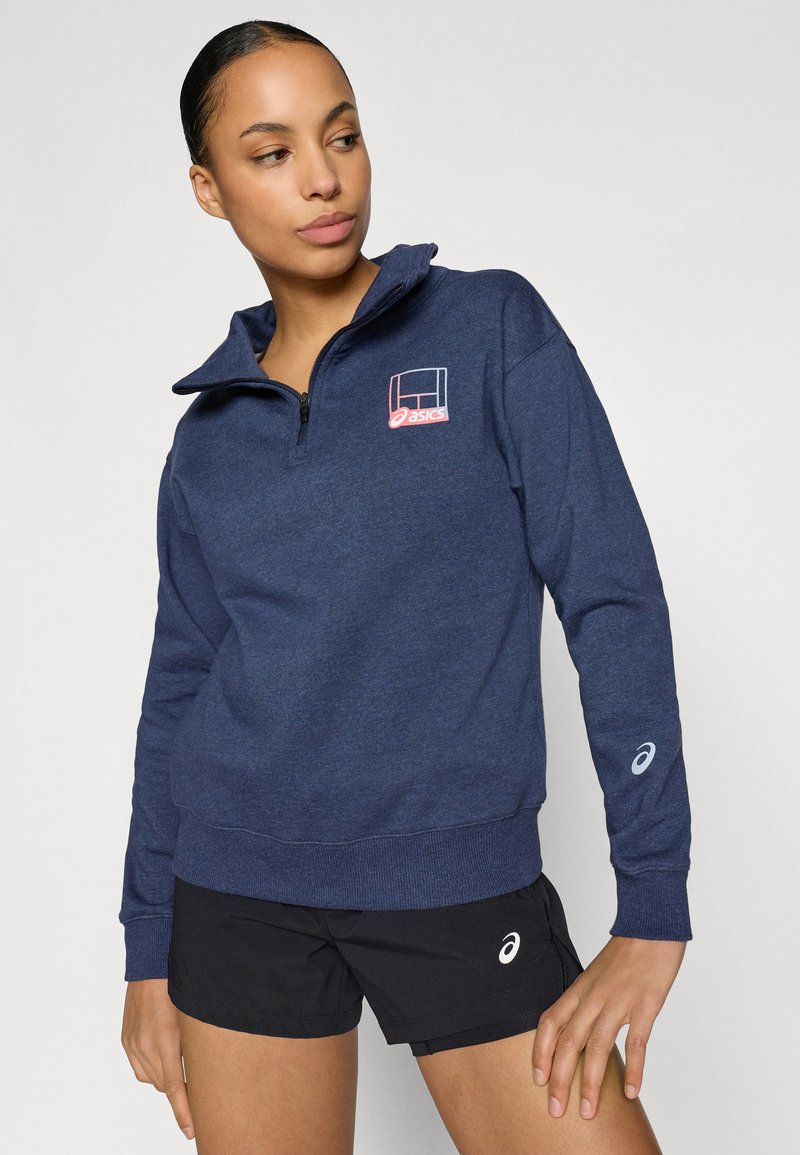 Donkerblauwe half-zip trui met een logo op de borst, geribbelde manchetten en een zachte stoftextuur. Gecombineerd met zwarte sportshorts.