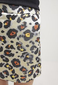 Close-up van een beige rok met zwart, oranje en gele luipaardprint, gedragen met een zwarte top tegen een effen grijze achtergrond.