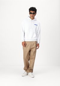 Felpa bianca con logo blu, pantaloni beige e sneakers bianche. Modello con occhiali da sole neri, in piedi contro uno sfondo neutro.