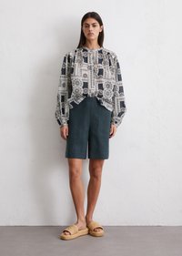 Geprinte blouse met pofmouwen, marineblauw en crème patroon; groene linnen shorts; geweven sandalen met een dikke zool. Neutrale achtergrond.