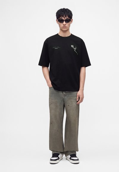 Νέος άνδρας με μαύρο γραφικό t-shirt, γκρι φαρδιά παντελόνια, μαύρα και άσπρα αθλητικά παπούτσια και σκούρα γυαλιά ηλίου, στέκεται με το ένα χέρι στην τσέπη.