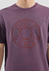 Fioletowa bawełniana koszulka z okrągłą grafiką zawierającą słowa "QUITE" i "SENSE" na czerwono. Tkanina ma gładką teksturę.