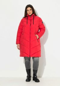 Cappotto imbottito rosso con cappuccio, caratterizzato da un motivo a zig zag, chiusura con zip e dettagli neri. Indossato sopra pantaloni grigi e stivali neri.