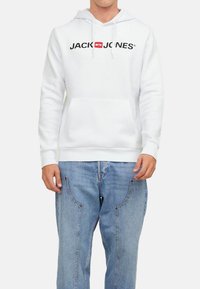 Hoodie blanc en coton avec une poche avant, arborant un logo noir 'JACK & JONES'. Associe à un jean en denim bleu clair.