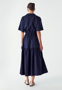 Tamsiai mėlyna, iki kelių siekianti suknelė su trumpomis rankovėmis, įtemptu juosmeniu ir sluoksniuota sijonu. Pagaminta iš minkštos medžiagos su lygiu paviršiumi. Matomos plokščios basutės.