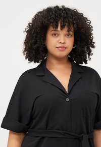 A young woman with curly hair wears a black collared outfit. She looks directly at the camera, exuding confidence and poise.