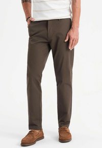Pantalons marron en tissu texturé, avec une coupe slim, accompagnés de chaussures en cuir marron. Lignes épurées et accents subtils.