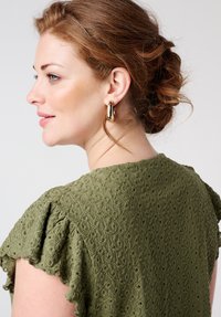 Haut texturé olive avec des manches volantées, présentant un tissu à motifs. De grandes boucles d'oreilles dorées ornent le profil du mannequin.