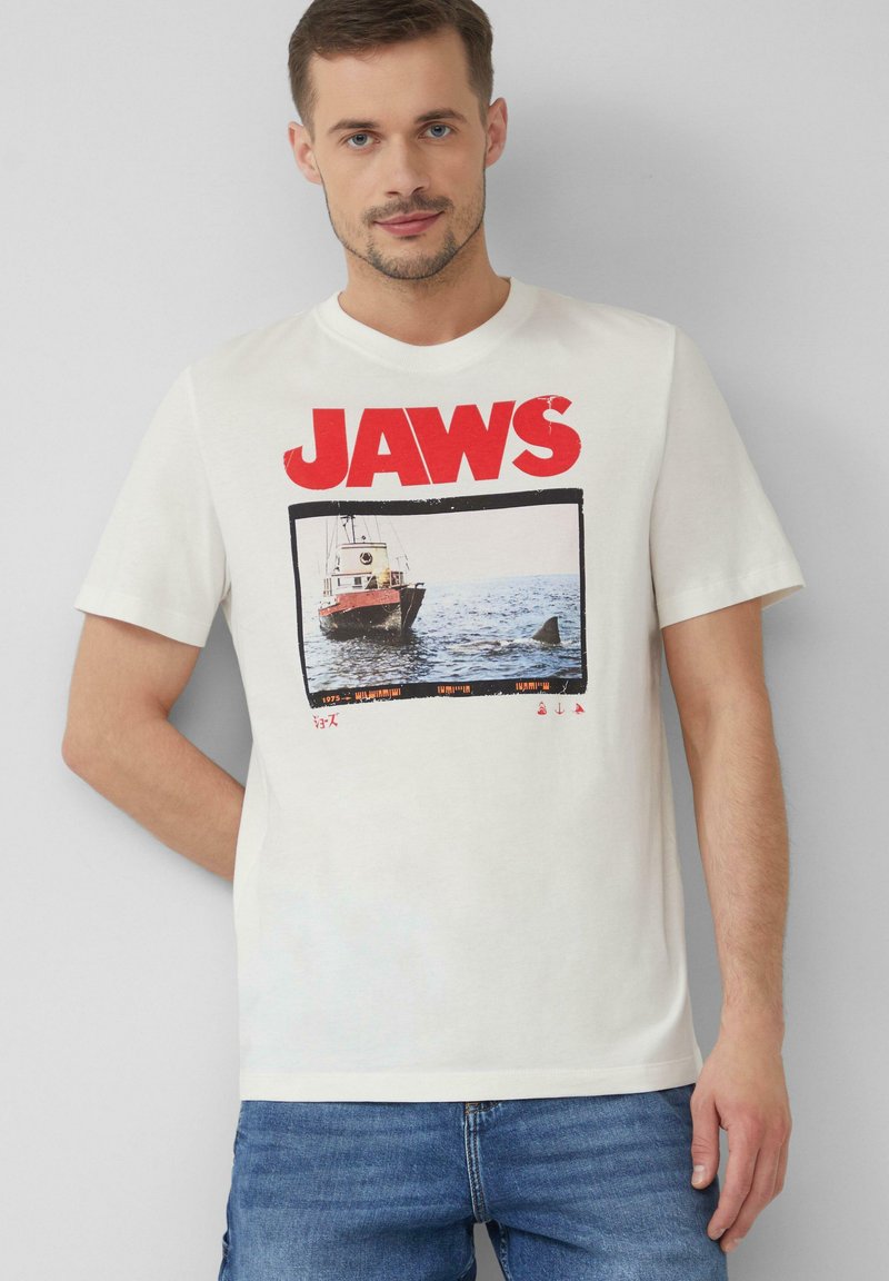Weißes Baumwoll-T-Shirt mit einem großen roten "JAWS"-Grafik und einem Filmstill von einem Boot und einem Hai, mit kurzen Ärmeln und einem Rundhalsausschnitt.