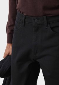Pantaloni in denim nero con un design aderente, dotati di tasche frontali, chiusura con bottone e dettagli di cucitura raffinati. Abbinati a un maglione scuro.