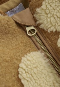 Nahaufnahme einer beigefarbenen und cremefarbenen strukturierten Wolljacke mit einem goldenen Reißverschluss und einem teilweise sichtbaren Stoffetikett im Kragen.