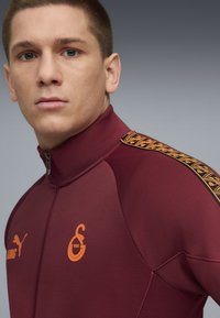 Vinsportjakke i bordeaux med høj krave, lynlås, orange logo og mønstrede detaljer på skuldrene i sort og orange.