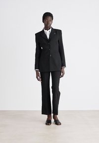 Blazer strutturato nero con rever a punta e bottoni, abbinato a pantaloni neri su misura e sandali neri aperti. Tessuto morbido.