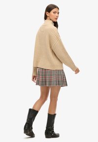 Un pull en maille côtelée beige avec un col montant, associé à une jupe à carreaux grise. Le modèle porte des bottes noires jusqu'aux genoux, affichant une silhouette décontractée.