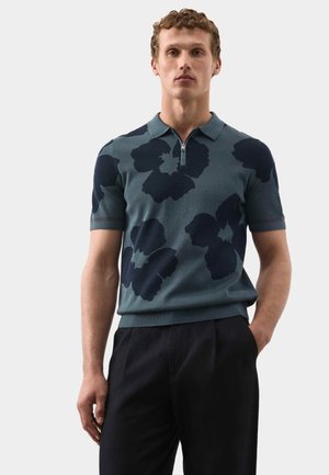 Uomo che indossa una polo con zip a maniche corte di colore blu-verde con grandi motivi floreali scuri, abbinata a pantaloni neri, in piedi davanti a uno sfondo semplice.