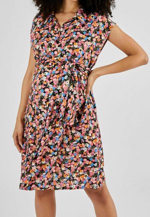 Kobieta w krótkiej, do kolan sukience w kwiaty z krótkimi rękawami i paskiem wiązanym w talii, stojąca z jedną ręką na biodrze.