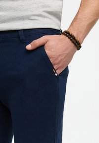 Pantaloni bleumarin cu un material texturat, buzunar lateral și o accente subtilă lângă buzunar. O brățară din mărgele maro este purtată pe încheietura mâinii.