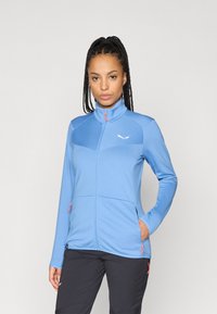 Ljusblå fleecejacka med hög krage, hel dragkedja, texturerad tyg och två fickor med dragkedja. Har orangea dragkedjor och logotyp.