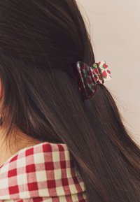 Dark brown hair held back with a translucent hair clip patterned with red strawberries, against a background of a red and white checkered collar.